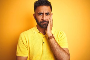Izole sarı arka plan üzerinde duran polo giyen genç hintli adam yorgun görünümlü ve çapraz kolları ile depresyon sorunları ile sıkılmış düşünme.