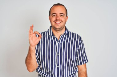 Izole beyaz arka plan üzerinde duran mavi çizgili gömlek giyen genç adam el ve parmakları ile ok işareti yapıyor olumlu gülümseyerek. Başarılı ifade.