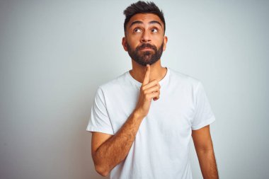İzole beyaz arka plan üzerinde duran t-shirt giyen genç hintli adam çene üzerinde parmak ile şüphe hakkında konsantre ve merak bakarak