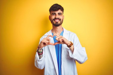 Dövme ile genç doktor adam izole sarı arka plan üzerinde duran aşk kalp sembolü ve elleriyle şekil gösteren gülümseyerek. Romantik kavram.