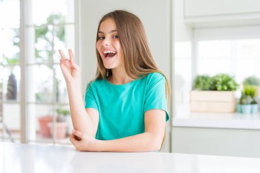 Yeşil tişört giyen güzel genç kız çocuk mutlu yüzüyle gülümseyerek zafer işareti yaparak kameraya göz kırpıyor. İki numara.