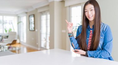 Uzun saçlı genç güzel asyalı kadın yüzünde büyük bir gülümseme ile kot ceket giyen, kameraya bakarak yan el parmağı ile işaret.