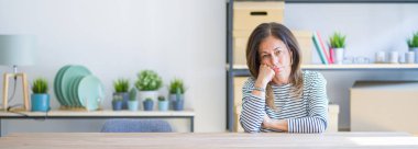 Orta yaş yaşlı kadın evde masada oturan geniş açılı fotoğraf yorgun görünümlü ve çapraz kolları ile depresyon sorunları ile sıkılmış düşünme.