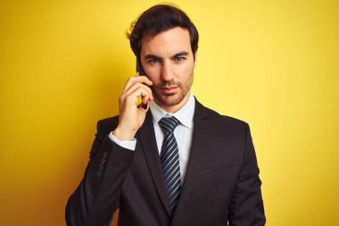 Young handsome businessman talking on the smartphone over isolated yellow background with a confident expression on smart face thinking serious