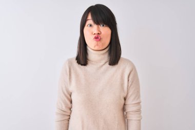 Genç güzel Çinli kadın komik yüzü ile izole beyaz arka plan üzerinde boğazlı kazak giyiyor yanaklar şişirme. Ağız hava ile şişirilmiş, deli ifade.