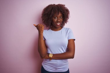 Genç afrikalı afro kadın izole pembe arka plan üzerinde duran t-shirt giyen mutlu yüz görünümlü gülümseyerek ve başparmak ile yan işaret.