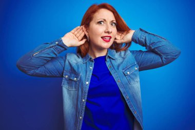 Mavi izole arka plan üzerinde duran kot gömlek giyen genç güzel kızıl saçlı kadın kulak jestiki elini duymak için çalışıyorum, dedikodu için meraklı. İşitme sorunu, sağır