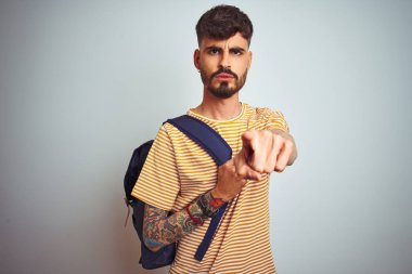 Kamera ve size parmak ile işaret izole beyaz arka plan üzerinde duran sırt çantası giyen dövme giyen genç öğrenci adam, el işareti, önden olumlu ve kendinden emin jest