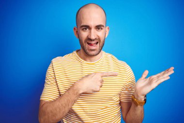 Sakallı genç kel adam mavi izole arka plan üzerinde rahat t-shirt giyen hayran ve el ile sunarken kameraya gülümseyerek ve parmak ile işaret.