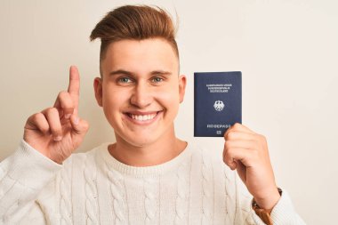 Izole beyaz arka plan üzerinde Almanya Alman pasaportu tutan genç yakışıklı adam mutlu yüz, bir numara ile bir fikir veya soru işaret parmağı ile şaşırttı