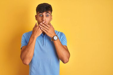 İzole sarı arka plan üzerinde duran mavi polo giyen genç hintli adam hata için elleri ile ağız kapsayan şok. Gizli kavram.