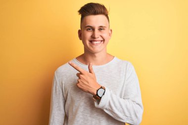 Genç yakışıklı adam beyaz rahat t-shirt giyen izole sarı arka plan neşeli yüzünde mutlu ve doğal ifade ile yan yana el ve parmak ile işaret eden yüz bir gülümseme ile neşeli