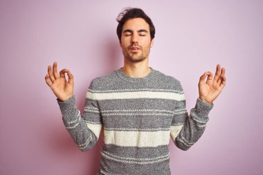 Genç yakışıklı adam izole pembe arka plan üzerinde duran rahat çizgili kazak giyen rahat ve gözleri parmakları ile meditasyon jest yapıyor kapalı gülümseyerek. Yoga konsepti.