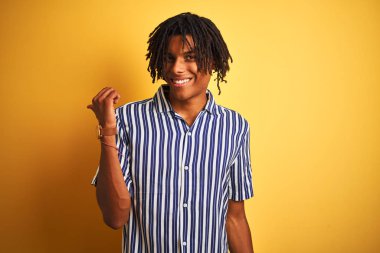 Dreadlocks ile rahat çizgili t-shirt izole sarı arka plan üzerinde mutlu yüz görünümlü gülümseyen giyen ve başparmak ile yan işaret afro adam.