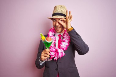 Takım elbise şapka hawaiian lei içme kokteyli giyen işadamı mutlu yüzü ile izole pembe arka plan üzerinde kokteyl içme parmakları na bakarak el ile ok işareti yapıyor gülümseyerek