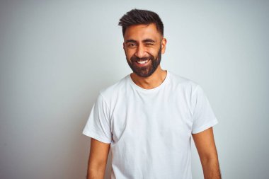 Genç hintli adam izole beyaz arka plan üzerinde duran t-shirt giyen seksi ifade, neşeli ve mutlu yüz ile kameraya bakarak göz kırpıyor.