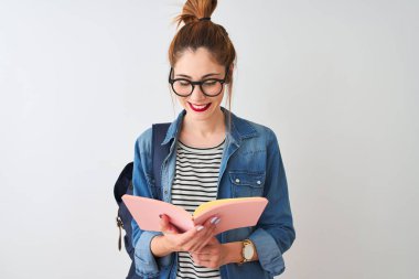 Redhead öğrenci kadın ayakta mutlu bir yüz ile izole beyaz arka plan üzerinde sırt çantası okuma kitabı giyen ve dişleri gösteren kendinden emin bir gülümseme ile gülümseyen