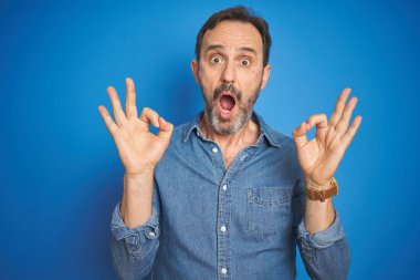 Izole mavi arka plan üzerinde gri saçlı Yakışıklı orta yaş yaşlı adam şaşırmış görünümlü ve parmakları ile ok onay sembolü yapıyor şok. Çılgın ifade