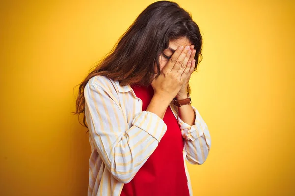 Young beautiful woman wearing red t-shirt and stripes shirt over yellow isolated background with sad expression covering face with hands while crying. Depression concept.