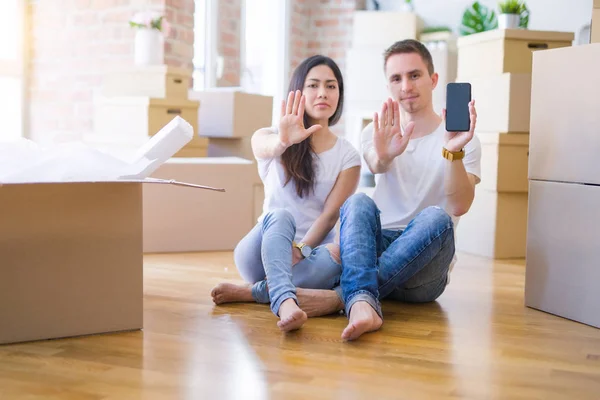 Beautiful couple sitting on the floor holding smartphone at new home around cardboard boxes with open hand doing stop sign with serious and confident expression, defense gesture