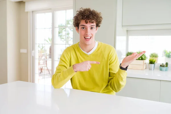 在家穿着黄色毛衣的年轻帅哥惊讶 微笑着对着镜头 一边用手和手指指着 — 图库照片