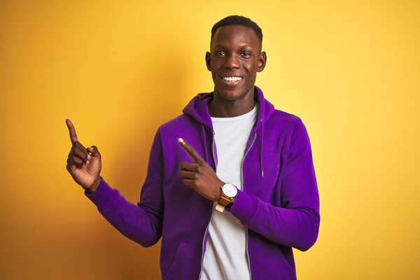 African american man wearing purple sweatshirt standing over isolated yellow background smiling and looking at the camera pointing with two hands and fingers to the side.