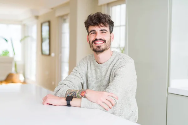 Young man wearing casual sweater sitting on white table happy face smiling with crossed arms looking at the camera. Positive person.