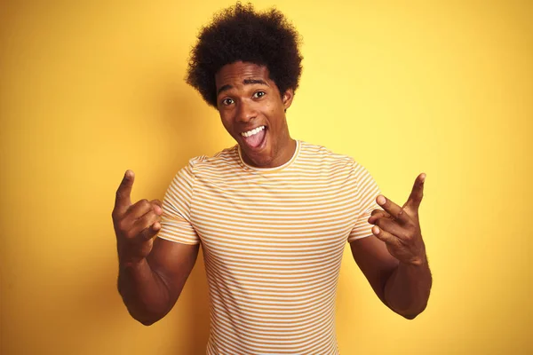American man with afro hair wearing striped t-shirt standing over isolated yellow background shouting with crazy expression doing rock symbol with hands up. Music star. Heavy music concept.
