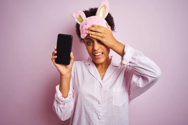 非洲妇女穿着睡衣和面具显示智能手机在孤立的粉红色背景强调与手在头上 震惊与耻辱和惊讶的脸 愤怒和沮丧 因错误而感到恐惧和不安 — 图库照片