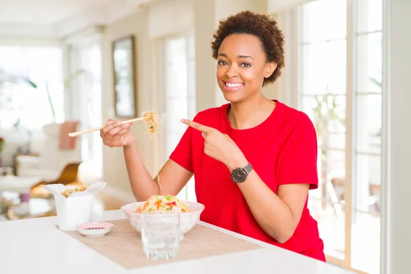 Young african american woman with afro hair eating asian food at home very happy pointing with hand and finger