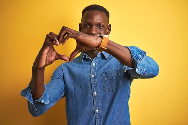 Young african american man wearing denim shirt standing over isolated yellow background smiling in love showing heart symbol and shape with hands. Romantic concept.