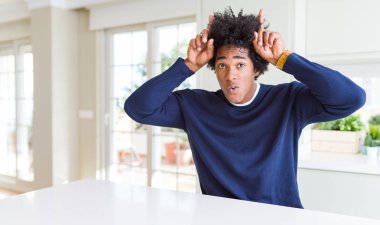 Genç afro-amerikan adam boğa boynuzları gibi baş üzerinde parmak ile komik jest yaparak evde oturan rahat kazak giyen