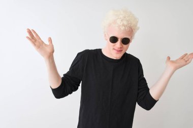 Genç albino adam siyah t-shirt giyen ve sunglasess izole beyaz arka plan üzerinde duran habersiz ve kolları ve elleri kaldırdı ile karışık ifade. Şüphe kavramı.