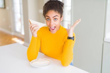 Genç afro-amerikan kadın sağlıklı atıştırmalık olarak bir sandviç yemek çok mutlu ve heyecanlı, kazanan ifade büyük bir gülümseme ile çığlık zafer kutluyor ve ellerini kaldırdı