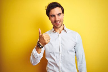 Genç yakışıklı işadamı zarif gömlek giyen izole sarı arka plan üzerinde duran el ile jest kadar mutlu başparmak yapıyor. Başarı gösteren kameraya bakarak ifadeyi onaylama.