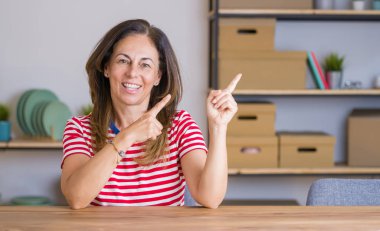 Orta yaş kıdemli kadın masada oturan evde gülümseyen ve iki el ve parmak tarafına işaret kamera bakıyor.