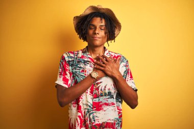 Izole sarı arka plan üzerinde çiçek gömlek ve şapka giyen dreadlocks ile Afro Amerikan adam kapalı gözleri ve yüzünde minnettar jest göğüs elleri ile gülümseyerek. Sağlık kavramı.