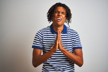 Çizgili mavi polo giyen dreadlocks giyen Afro adam izole beyaz arka plan üzerinde ayakta yalvarıyor ve çok duygusal ve endişeli yüzünde umut ifadesi ile birlikte elleri ile dua. Af dilemek istiyorum. Din kavramı.