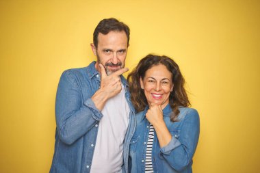 Güzel orta yaş çift birlikte izole sarı arka plan üzerinde kot gömlek giyen çapraz kollar ve el çene kaldırdı ile gülümseme ile kameraya emin görünümlü. Olumlu düşünme.