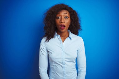 Izole mavi arka plan üzerinde duran gömlek giyen Afro-Amerikan iş kadını korkmuş ve sürpriz ifade, korku ve heyecanlı yüz ile şok.