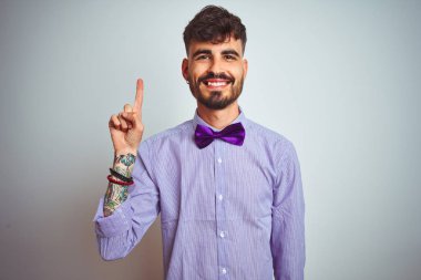 Dövme mor gömlek ve izole beyaz arka plan üzerinde papyon giyen genç adam gösteren ve kendinden emin ve mutlu gülümserken parmak sayısı bir işaret.