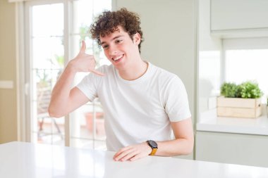 Evde beyaz tişört giyen genç yakışıklı adam telefonda konuşmak gibi el ve parmaklarla telefon hareketi yaparak gülümsüyor. İletişim kavramları.