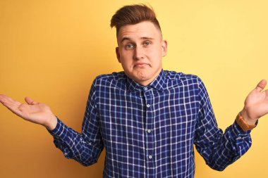 İzole edilmiş sarı arka planda dikilen sıradan tişört giyen genç yakışıklı bir adam. Kolları ve elleri havada, kafası karışmış bir ifade. Şüphe kavramı.