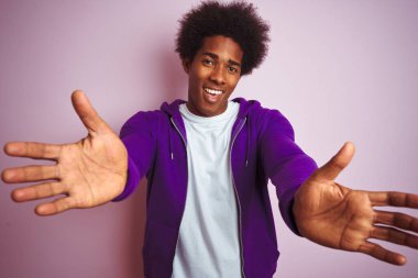 Mor sweatshirt giyen genç afro-amerikan adam izole pembe arka plan üzerinde duran sarılmak için açık kollarla gülümseyerek kameraya bakıyor. Mutluluğu kucaklayan neşeli ifade.