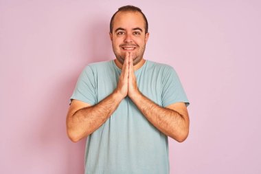 Mavi rahat t-shirt giyen genç adam emin gülümseyerek birlikte elleri ile dua izole pembe arka plan üzerinde duran.
