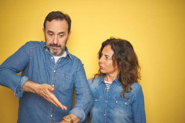 Güzel orta yaş çift birlikte izole sarı arka plan üzerinde duran acele zaman izlemek için işaret, sabırsızlık, üzgün ve son gecikme için kızgın