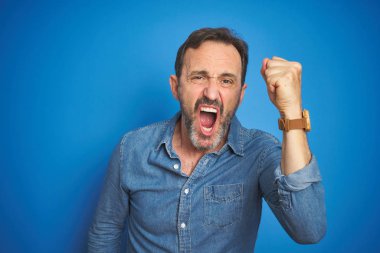 Izole mavi arka plan üzerinde gri saçlı Yakışıklı orta yaş yaşlı adam kızgın ve öfke ile bağırırken öfkeli ve öfkeli yumruğa yükselterek. Öfke ve agresif kavram.