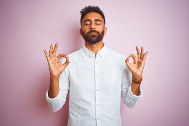 İzole pembe arka plan üzerinde duran zarif gömlek giyen genç Hintli işadamı rahatlayın ve parmakları ile meditasyon hareketi yaparken kapalı gözleri ile gülümseyerek. Yoga konsepti.