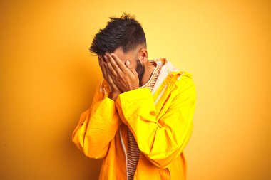 Genç hintli adam yağmurluk giyen izole sarı arka plan üzerinde ayakta üzgün ifade ile elleri ile yüz kapsayan ağlarken. Depresyon kavramı.