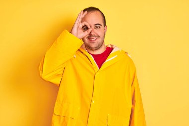 Izole sarı arka plan üzerinde duran yağmurluk giyen genç adam el gülümseyerek ok jest yapıyor, mutlu yüz ile parmaklardan bakan göz.